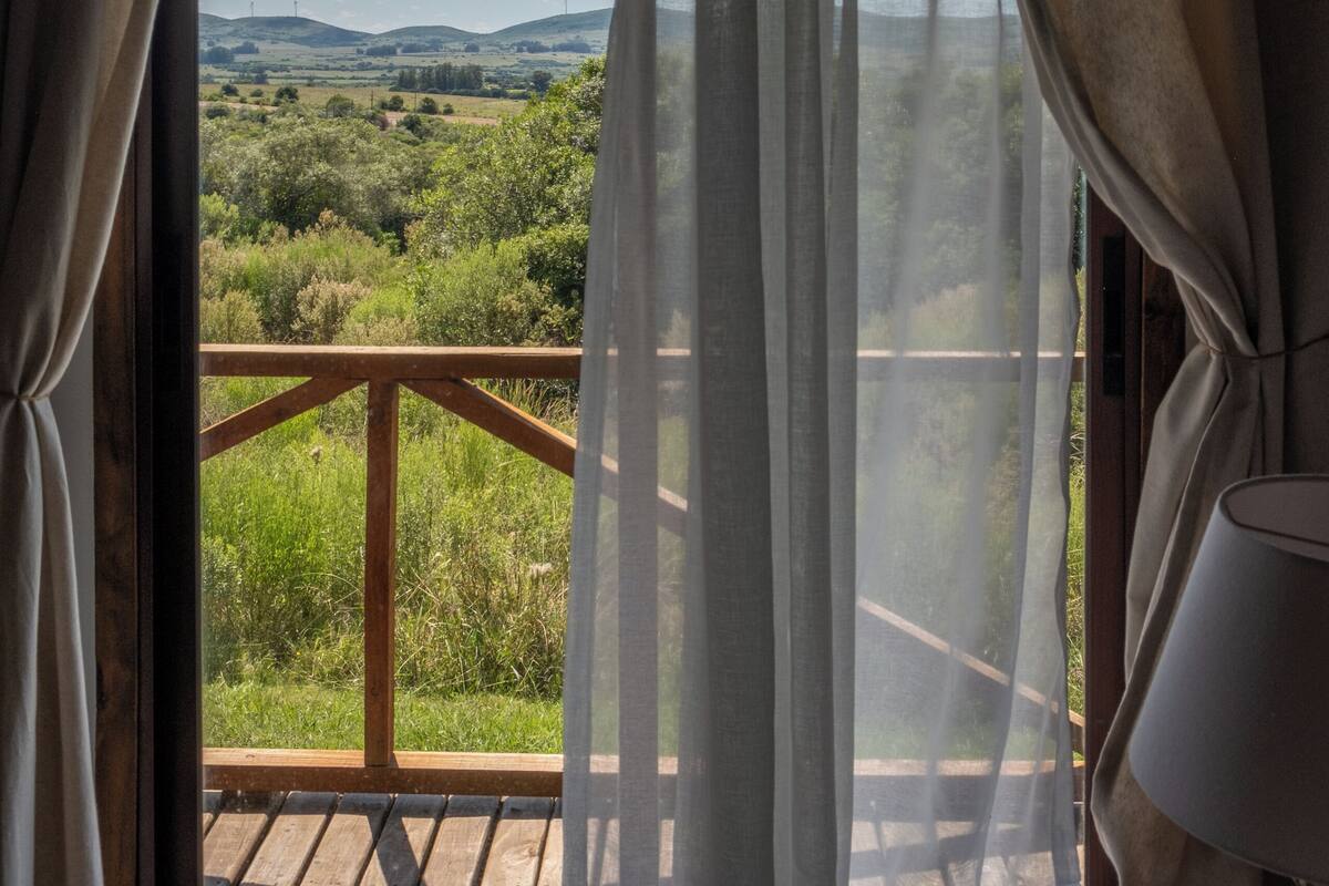 A room with a wooden balcony, a window with sheer curtains, and a lamp on the right side.