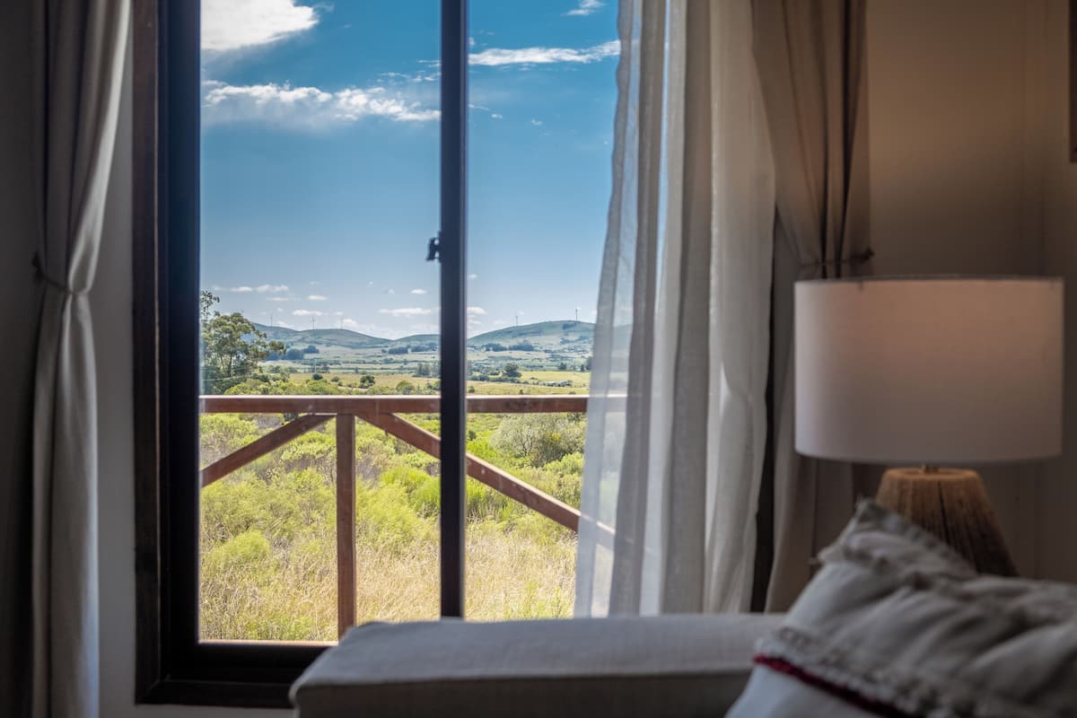 A room with a large window, a wooden railing, and a lamp on a bedside table.