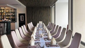 Une longue table à manger, dressée pour un repas formel, entourée de nombreuses chaises, un coin bar avec des bouteilles et un design de plafond moderne.
