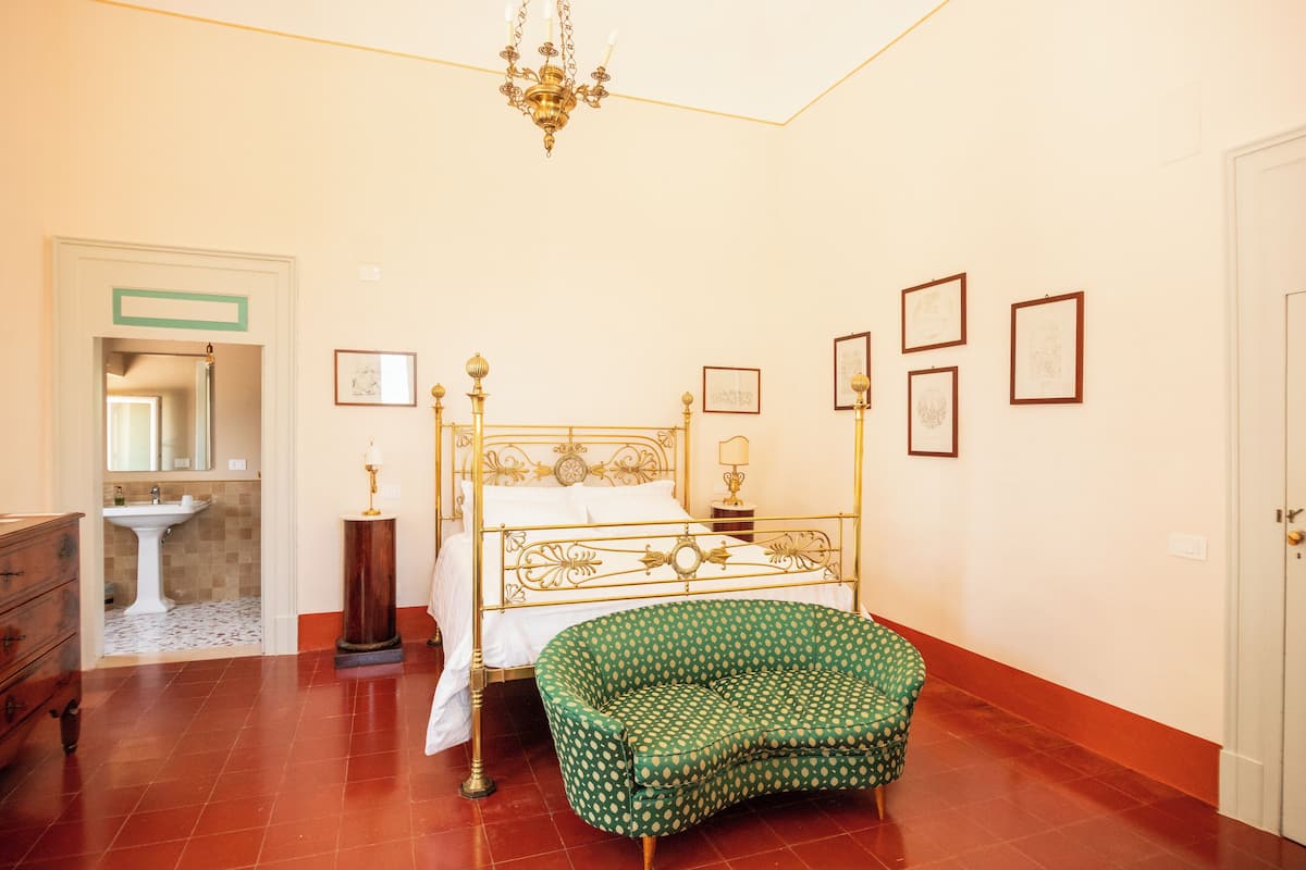 Ein Schlafzimmer mit einem Himmelbett, einem grün gemusterten Sessel, einer Kristallleuchtre und gerahmten Bildern an der Wand.