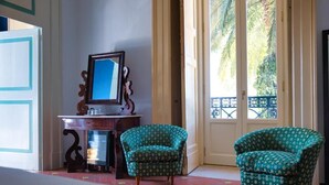 A room with a wooden desk, two patterned armchairs, a mirror, and a view of a balcony with a blue railing.