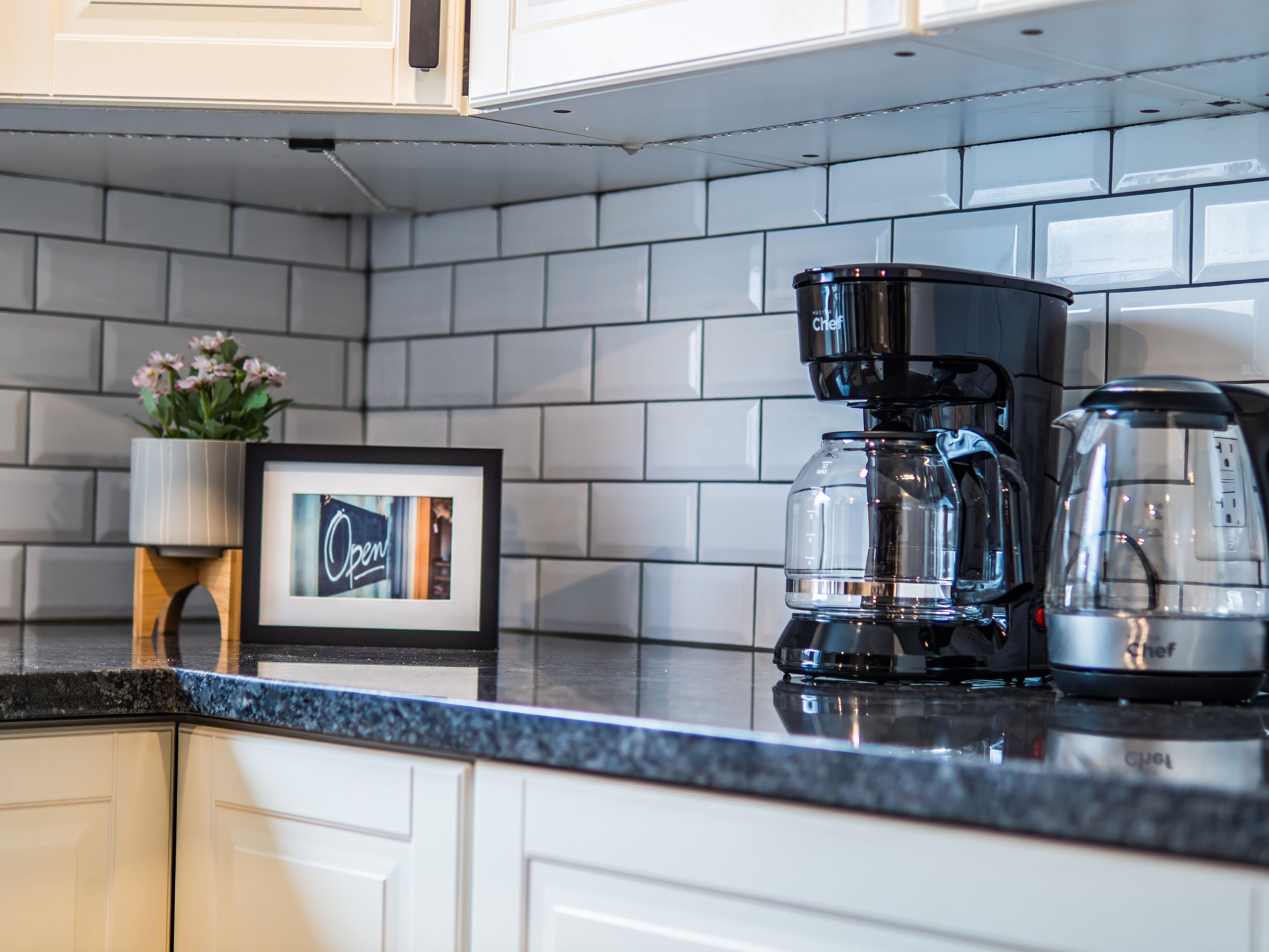 A kitchen with white cabinets, a black coffee maker, a glass carafe, and a framed picture on the counter.