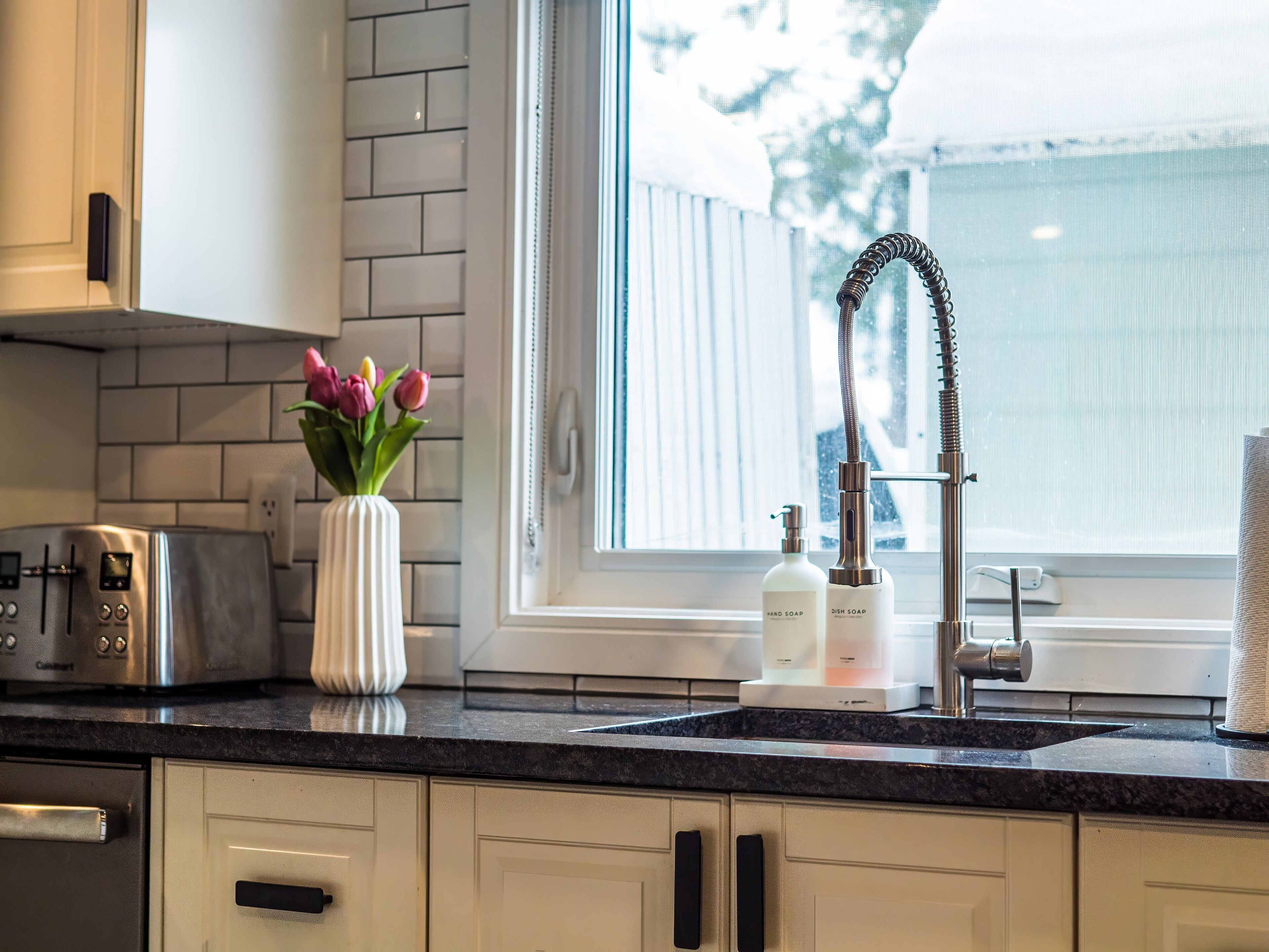 Eine Küche mit modernem Design, inklusive eines Edelstahltoasters, einer weißen Vase mit Blumen sowie einem Wasserhahn mit flexiblem Schlauch.
