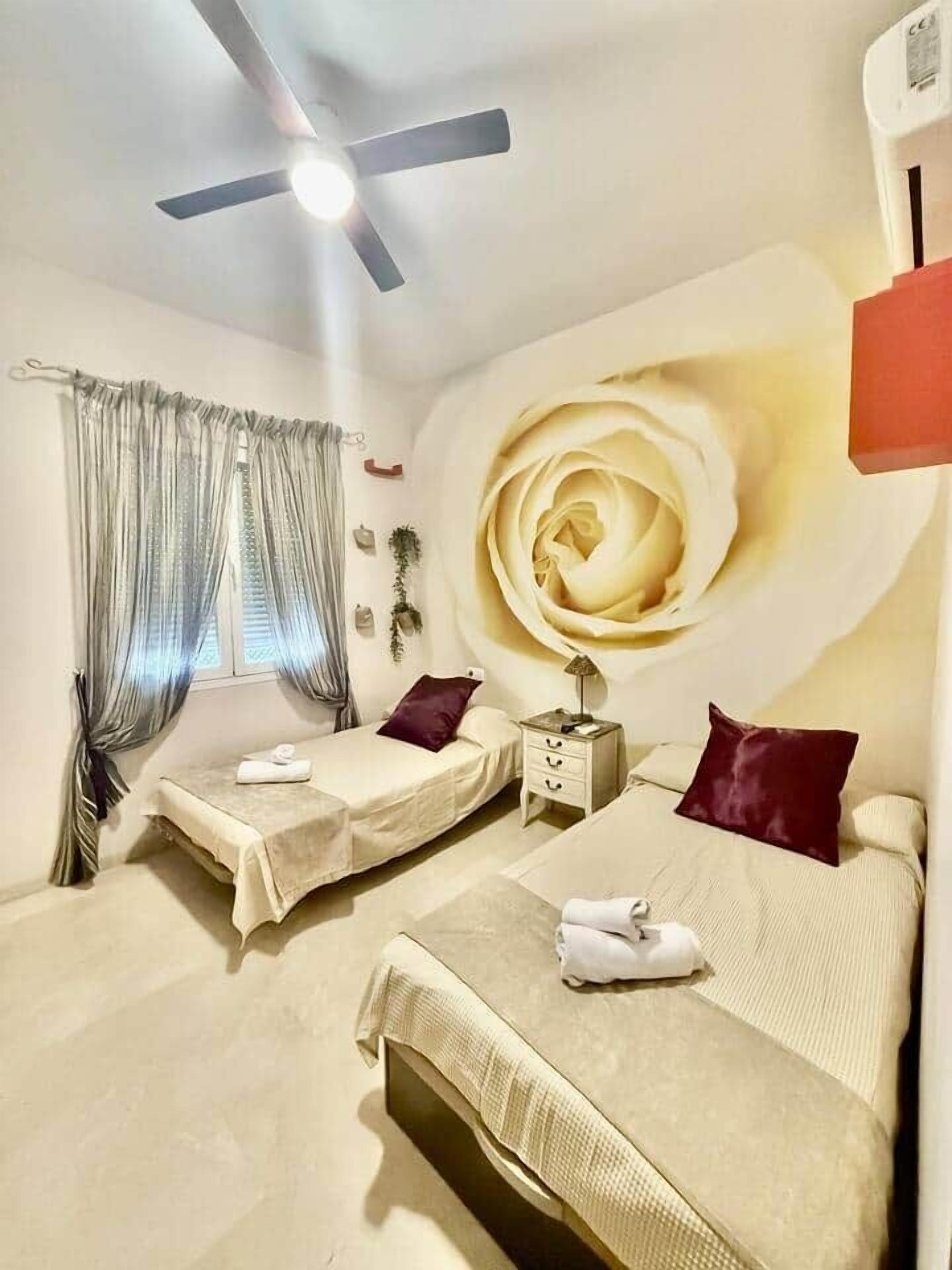 Een hotelkamer met twee bedden, een plafondventilator, een groot muurschildering van een roos en een raam met gordijnen.