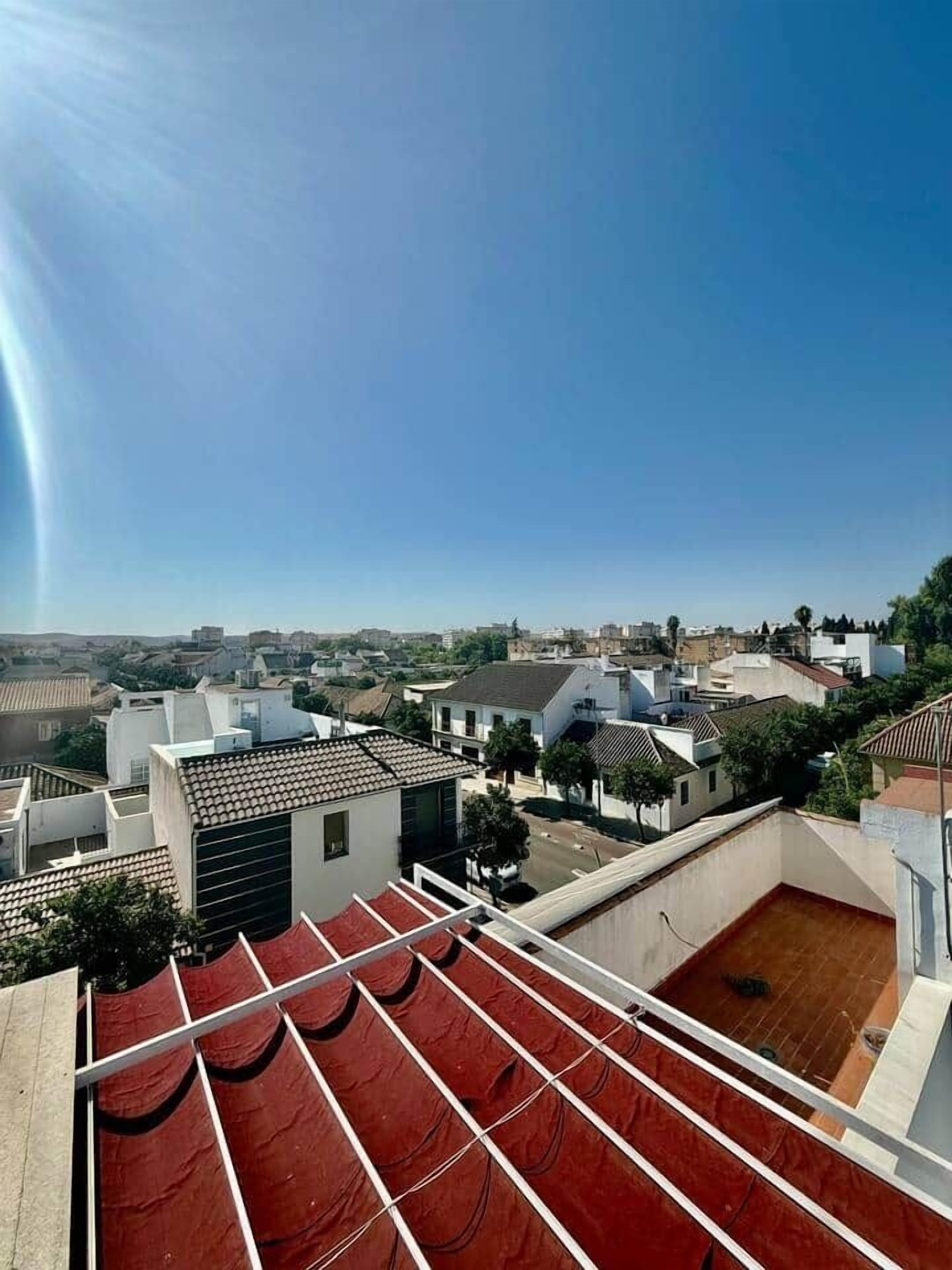 A rooftop terrace with red awnings, a view of a residential area with various buildings, and a clear blue sky.
