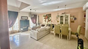 Un salon spacieux avec un canapé blanc, une table à manger et des chaises. On y trouve également un téléviseur, un lustre et une grande fenêtre avec des rideaux.
