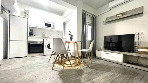 Cozinha moderna com armários brancos, forno embutido e micro-ondas. Há uma área de jantar com mesa redonda e cadeiras, e uma TV de tela plana na parede.