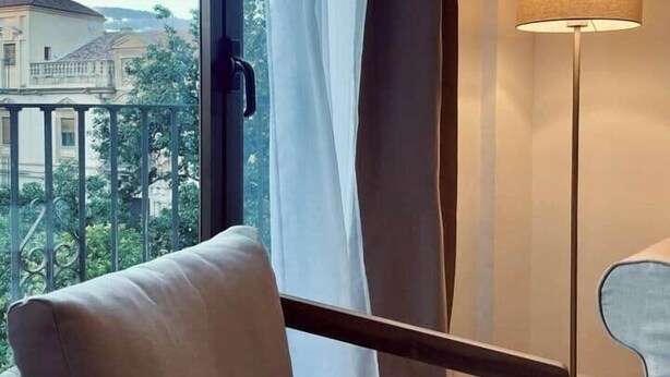Une chambre d’hôtel avec une grande fenêtre, un canapé blanc, un lampadaire et une vue sur les bâtiments et la verdure environnante.