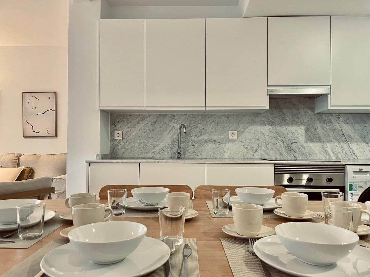 Cozinha moderna com armários brancos, painel de mármore e mesa de jantar posta para seis, com pratos, tigelas e copos brancos.