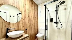 Un baño con tocador de madera, un espejo redondo, un inodoro y ducha con cerramiento de vidrio.