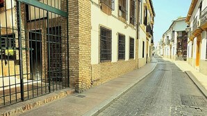 A narrow cobblestone street lined with buildings, featuring a brick wall with barred windows and a metal gate on the left. - Apartment in Casco Historico (Córdoba)