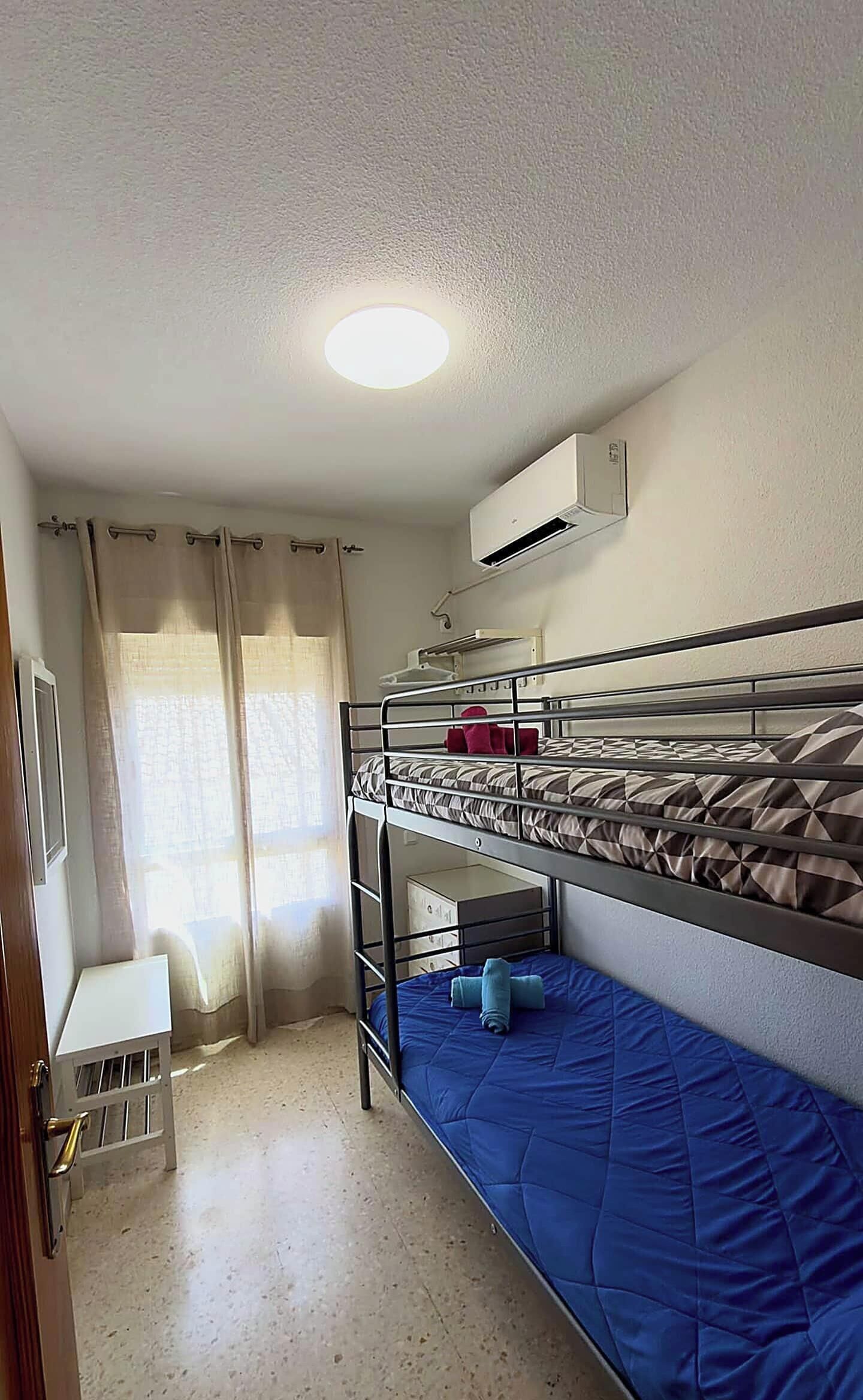 Une chambre avec un lit superposé, un ventilateur de plafond, une petite table et une fenêtre avec des rideaux.