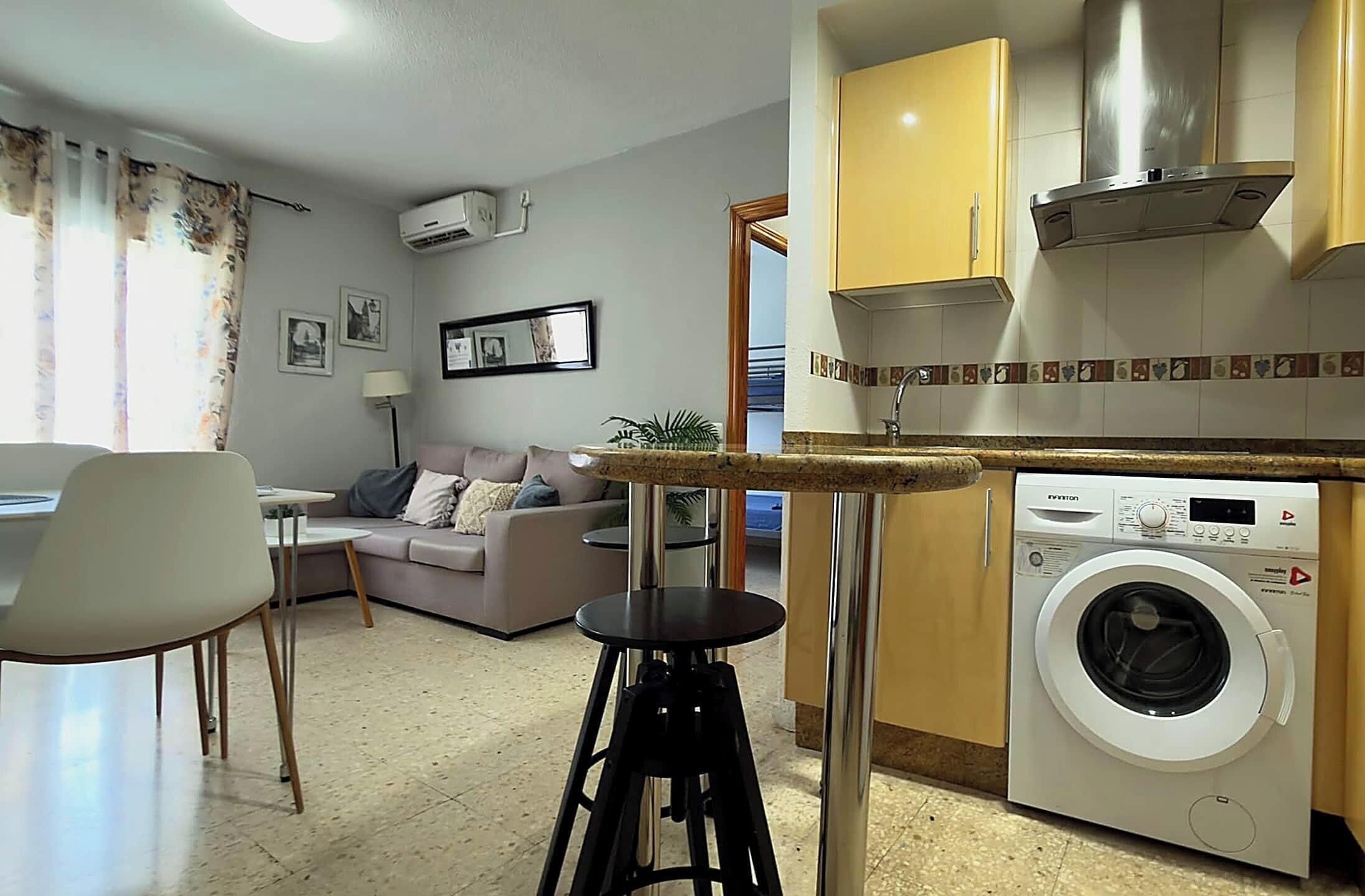 Un espace de vie compact comprenant un coin cuisine, un lave-linge, un canapé, un coin repas et une petite table.