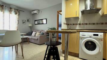 Un espace de vie compact comprenant un coin cuisine, un lave-linge, un canapé, un coin repas et une petite table.