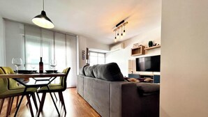 Een moderne woonkamer met een grijze bank, een eethoek met een tafel en stoelen, en een aan de muur bevestigde televisie.