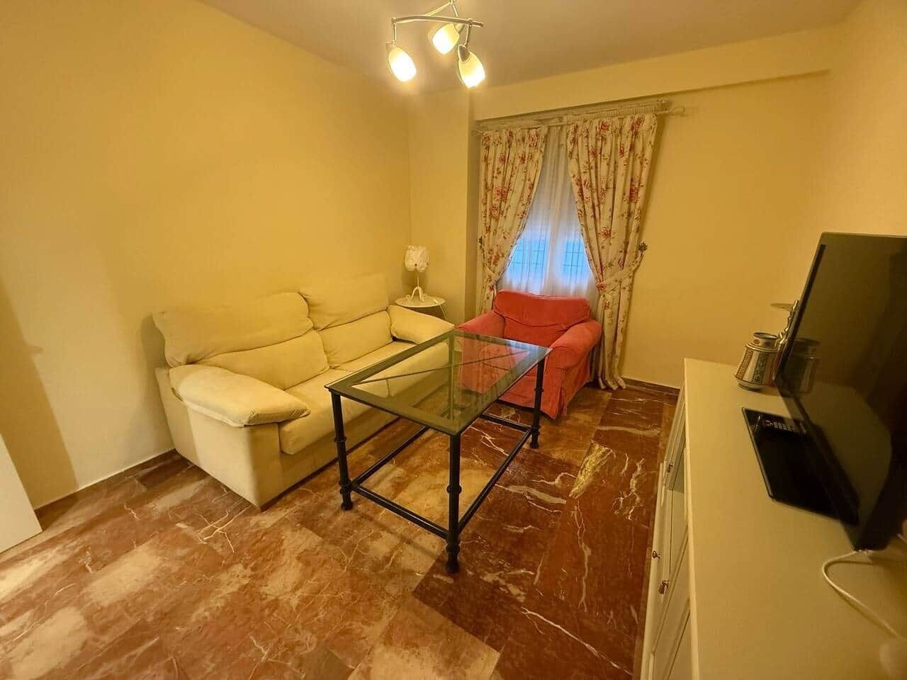Uma sala de estar com sofá, mesa de centro de vidro, uma poltrona vermelha, televisão e luminária.