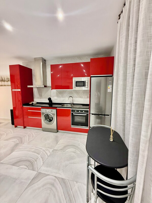 A modern kitchen with red cabinetry, stainless steel appliances, a washing machine, and a dining area with a table and chairs.