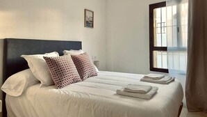 Un lit bien fait, avec du linge de lit blanc et des coussins à motifs, un petit tableau encadré accroché au mur et une fenêtre avec des rideaux.