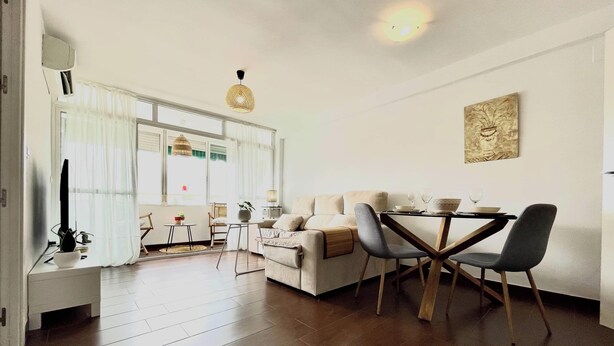 Uma sala de estar moderna com sofá, mesa de jantar e cadeiras. Há uma janela com cortinas, um lustre e um quadro na parede.