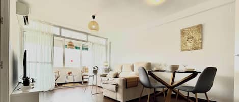 A modern living room with a sofa, dining table, and chairs. There is a window with curtains, a hanging light fixture, and a painting on the wall.