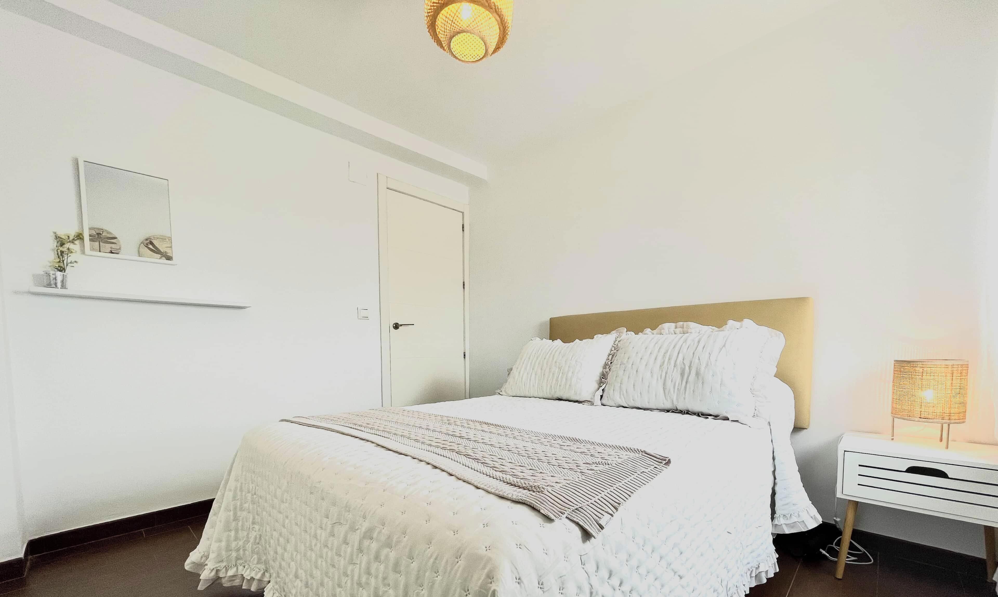 A bedroom with a bed, a bedside table, a lamp, a door, and a wall-mounted shelf with a plant.