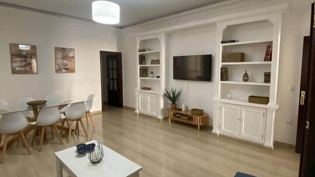 Ein modernes Wohnzimmer mit einem weißen Sofa, einem Esstisch mit Stühlen, einem Couchtisch, einem an einem weißen Schrank befestigten Fernseher und einer Deckenleuchte.