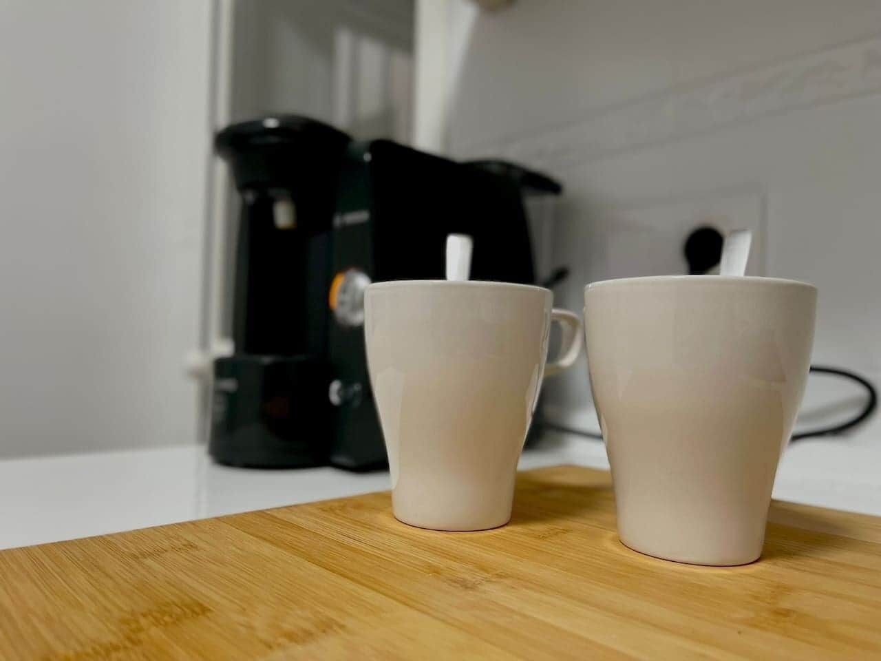 Deux tasses en céramique blanche sont posées sur une surface en bois, avec une machine à café en arrière-plan.