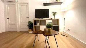 Une pièce avec un plancher en bois, une table à manger avec des chaises, un téléviseur fixé au mur, une bibliothèque et une lampe.