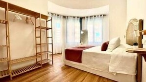 Una camera d'albergo con pavimento in legno, un letto con lenzuola bianche, una coperta rossa, un armadio in legno con abiti appesi e una finestra con tende.