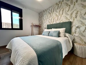 Ein Schlafzimmer mit Bett, Kopfstütze, Fenster und einer Wand mit Blattmuster, sowie ein kleiner runder Tisch.