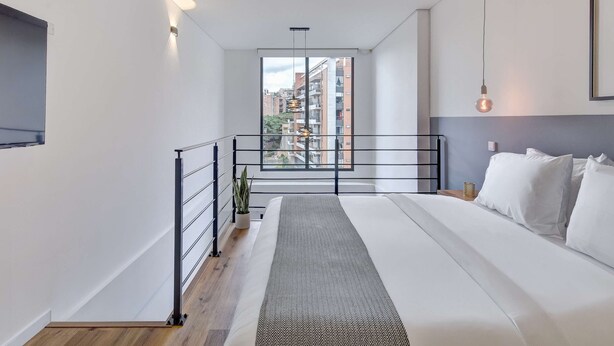 A modern bedroom with a large bed, a view of the city through a window, and a balcony with a railing. - w* |  Stylish & Cozy Duplex Loft in Zona G (Bogotá)