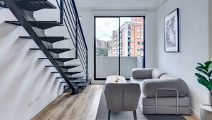 A modern living room with a staircase, a sofa, a coffee table, and a view of the city through a large window. - w* |  Stylish & Cozy Duplex Loft in Zona G (Bogotá)