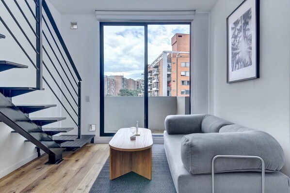 En modern lägenhet med en trappa, ett runt soffbord i trä, en grå soffa och ett stort fönster med utsikt över staden.
