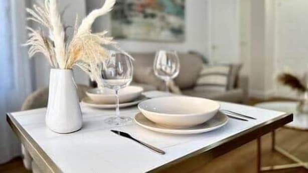 Uma mesa de jantar posta com pratos brancos, um vaso com plumas de capim-do-campo e taças de vinho.
