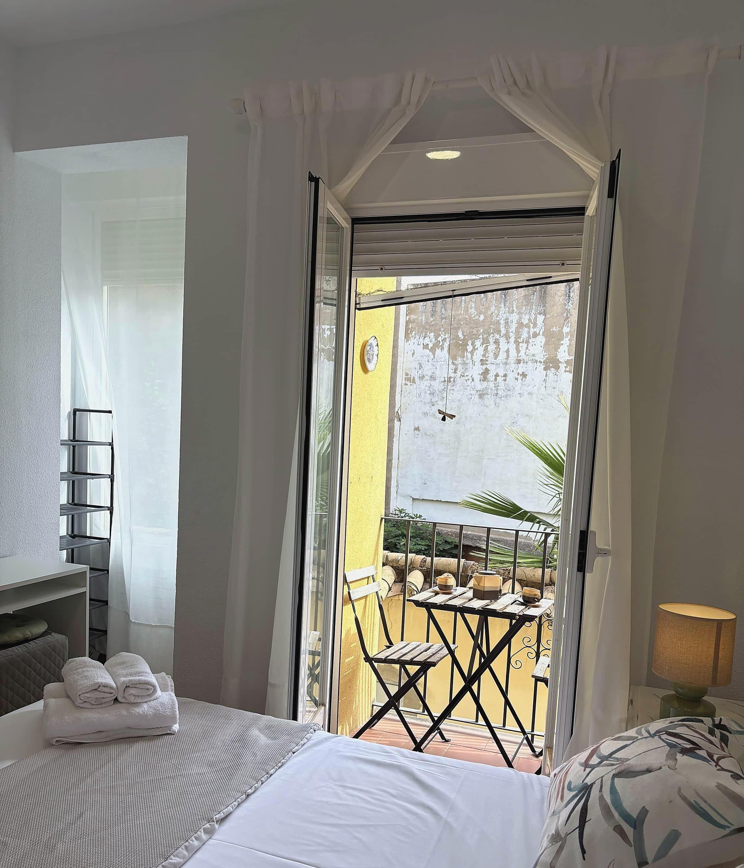 A hotel room with a bed, a small table with a lamp, and a balcony with a table and chairs.