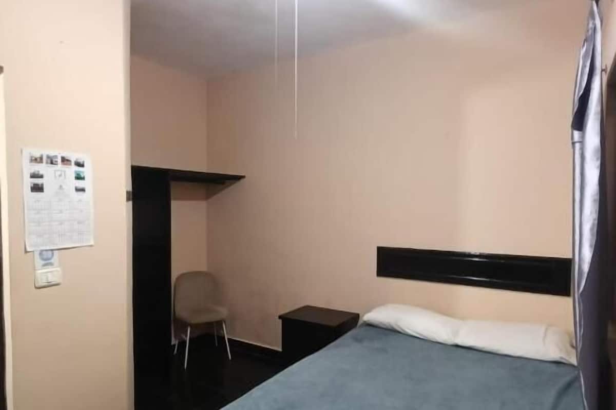 Une chambre d’hôtel équipée d’un lit, d’une chaise, d’une petite table, d’un ventilateur de plafond et d’un téléviseur fixé au mur.