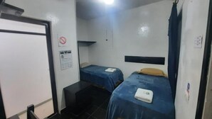 Una habitación pequeña y funcional con dos camas individuales, un ventilador de techo, una pizarra y una puerta.