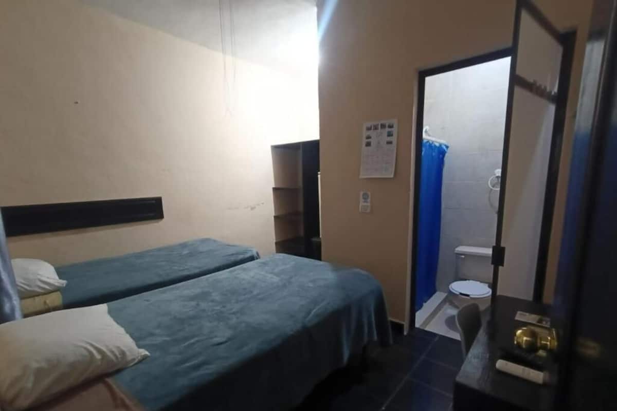 Une chambre d’hôtel avec deux lits simples, une petite table, un miroir et une salle de bain visible à travers une porte ouverte.