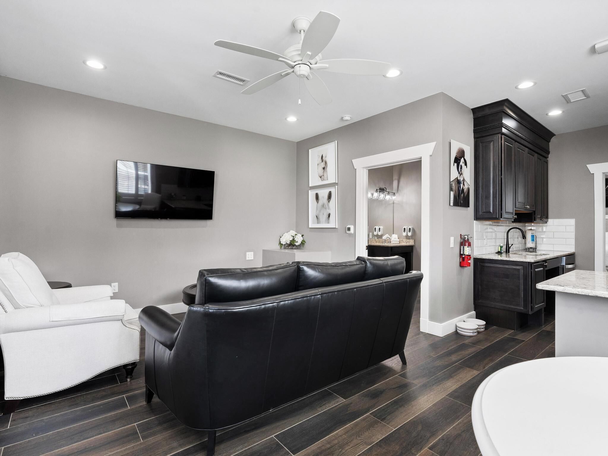 Una sala de estar moderna con un sofá de cuero negro, un sillón blanco, un televisor de pantalla plana y un ventilador de techo.