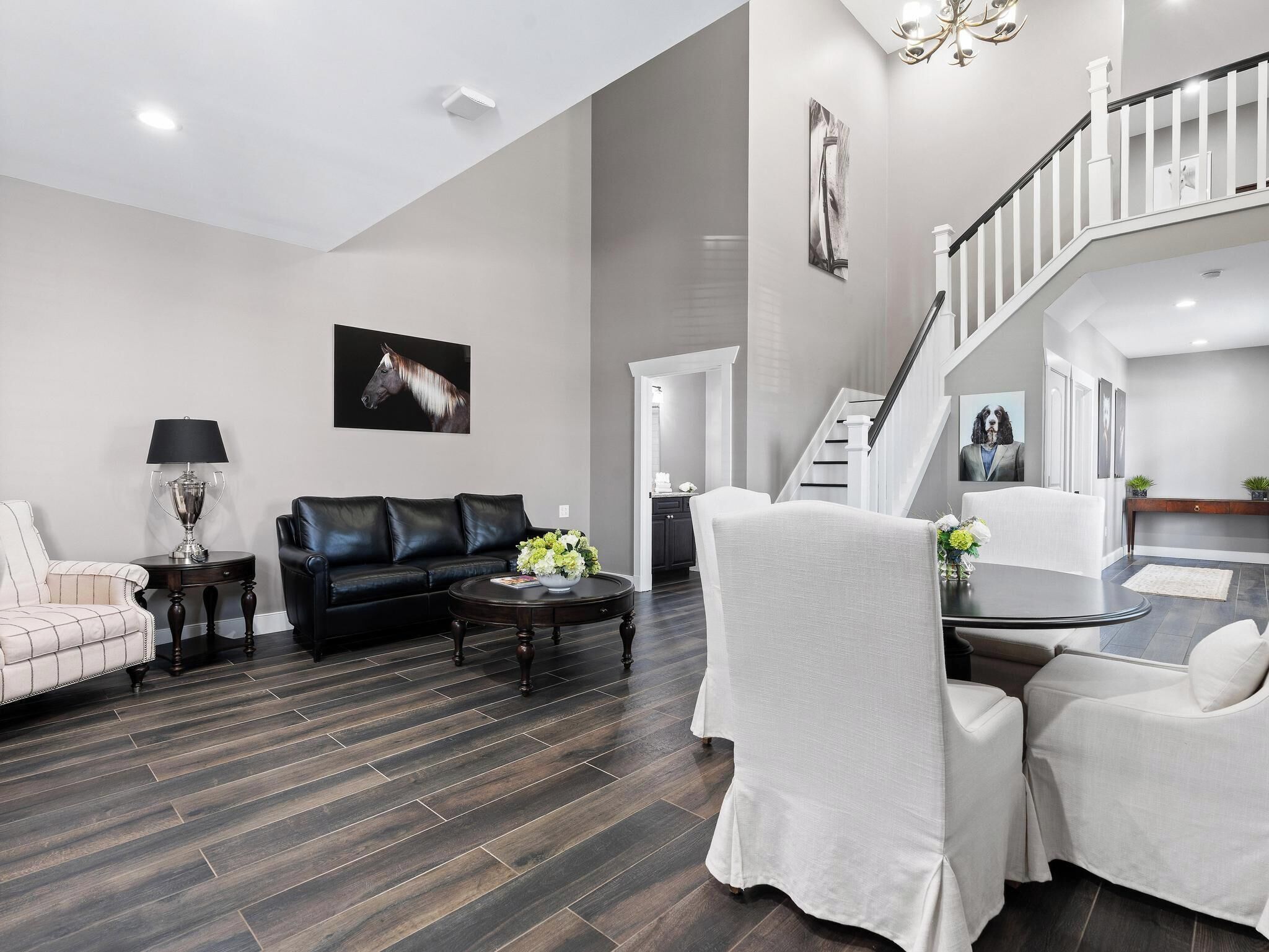 A modern living room with a black leather sofa, a white armchair, a coffee table, and a dining table with chairs. There is a staircase with a railing leading to an upper level.