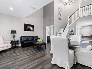 A modern living room with a black leather sofa, a white armchair, a coffee table, and a dining table with chairs. There is a staircase with a railing leading to an upper level. - World Equestrian Center Wilmington (Wilmington)