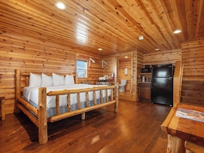 A log cabin interior with a wooden bed, a kitchen area, and a bathroom.
