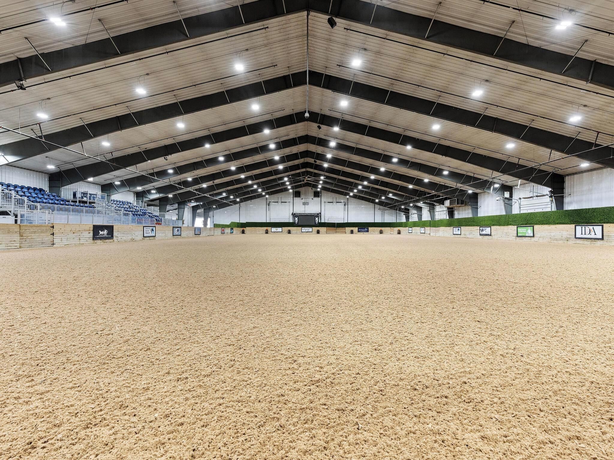 An indoor equestrian arena with a sandy floor, high ceilings, and seating areas.