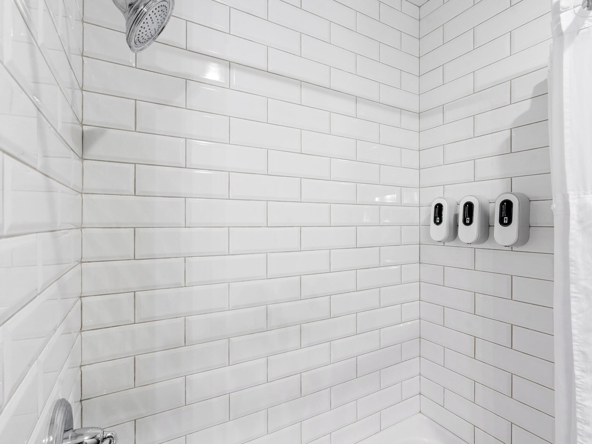 A white-tiled shower with a handheld showerhead and three soap dispensers.