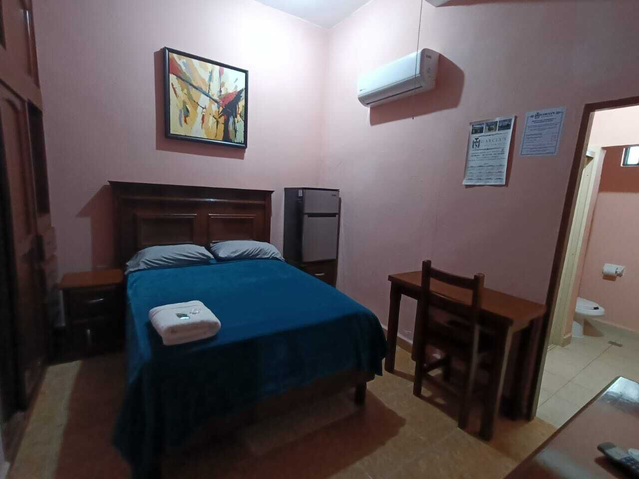 A bedroom with a bed, a desk, a chair, an air conditioner, and a painting on the wall.