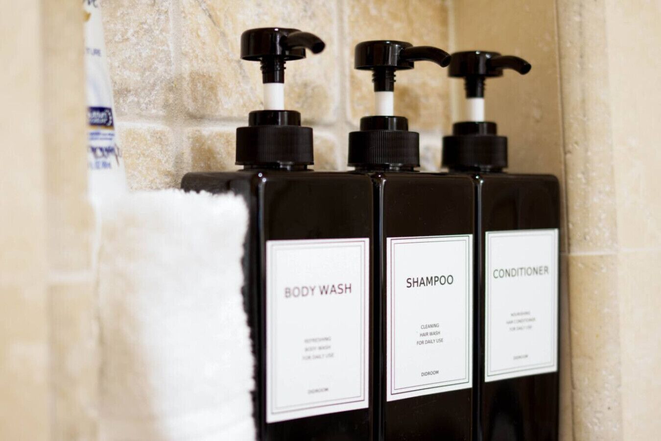 Kolme mustaa pulloa, joissa on pumppumekanismi ja joihin on merkitty tekstit ”KAPPALPESU”, ”SHAMPOO” ja ”HOITOAINE”, ja ne on asetettu laatoitetun seinän eteen.