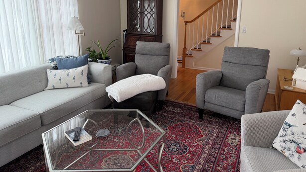 Ein Wohnzimmer mit grauem Sofa, zwei Sesseln, einem Glastisch, einer dunklen Holzkommode, einem roten Teppich mit Muster und einer Treppe, die zu einer oberen Etage führt.