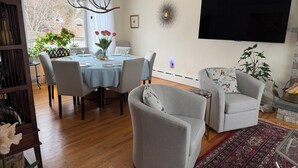 Ein Essbereich mit einem Tisch für vier Personen, umgeben von vier Stühlen. Über dem Tisch hängt eine Chandelieber, an der Wand ist ein Flachbildfernseher angebracht und auf dem Fensterbrett steht ein Blumentopf.