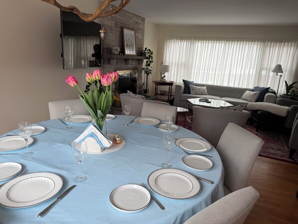 A dining area with a round table set for six, surrounded by chairs. There is a vase with flowers on the table, and a fireplace with a stone mantle in the background.
