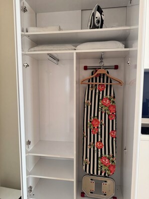 A white wardrobe with multiple shelves, a hanging rod, and a small ironing board with a floral iron. - Orbi City Aparts room (Batumi)